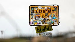 Das Ortsschild "Lützerath" ist beklebt mit bunten Stickern. Unterhalb des Schilds wurde ein Holzschild mit der Aufschrift "bleibt!" angebracht.