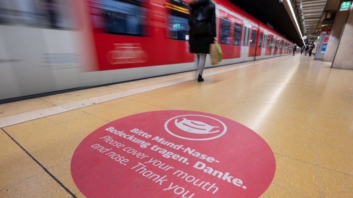 Ein Aufkleber weist in einer S-Bahn Haltestelle auf das Tragen einer Mund-Nasen-Bedeckung hin. Im Hintergrund fährt ein Zug vorbei.