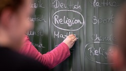 Eine Lehrerin schreibt mit Kreide einen Stundenplan mit Religionsunterricht an eine Tafel.