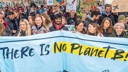 Eine Gruppe von Aktivist:innen steht zusammen und hält ein langes Plakat mit der Aufschrift "There is no Planet B!" in der Hand. Im Hintergrund sind weitere Demonstrant:innen zu sehen, die verschiedene selbstbemalte Plakate tragen.