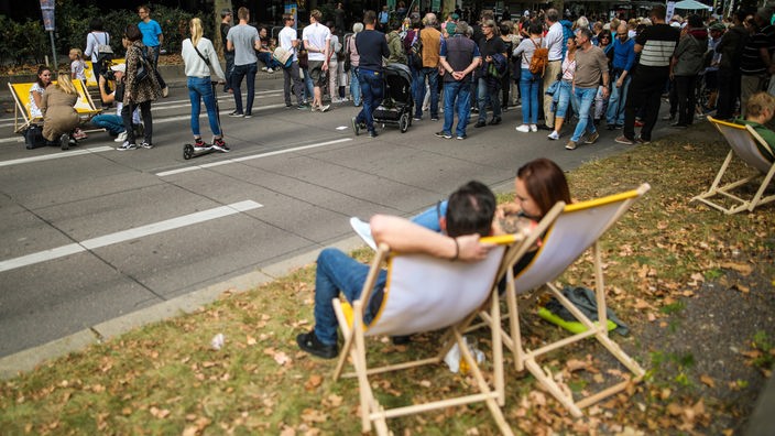 Menschen laufen über die für den Autoverkehr gesperrte Theodor-Heuss-Straße in der Stuttgarter Innenstadt. Zwei Personen sitzen in Liegestülen.