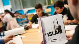 Ein Schüler öffnet in einem Klassenraum einen Schnellhefter mit einem selbst gestalteteten Deckblatt für das Fach Islamunterricht.