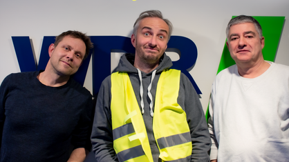 Henning Bornemann, Jan Böhmermann in gelber Warnweste und Axel Naumer posieren locker vor dem WDR5-Logo auf dem Flur der WDR-Arkaden