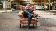 Der ganz in schwarz gekleidete Bülent Ceylan sitzt in einer alten und heruntergekommenen Industriehalle in einem ledernden Ohrensessel, Foto: Daniel Haussmann
