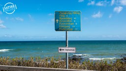 Bildmontage Wegweiser: Eine Tafel mit griechischen Städtenamen weist aufs Meer, zum Impfzentrum geht es links entlang