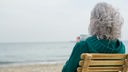 Grauhaarige Frau sitzt mit einer Tasse Tee auf einem Bambusstuhl am Strand 