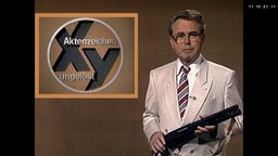 Eduard Zimmermann steht vor einem braunen Hintergrund mit dem Sendungslogo "Aktenzeichen XY ... ungelöst"