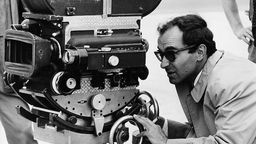 Schwarzweiß-Foto von Jean-Luc Godard, der bei Dreharbeiten durch eine Kamera blickt.
