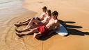 Drei Jugendliche blicken am Strand, auf ein Surfbrett gestützt, ins Sonnenlicht aufs Wasser.