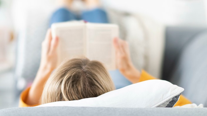 Symbolbild: Eine junge Frau liegt auf einem Sofa und liest ein Buch.