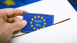 Hand wirft eine Europa-Fahne mit Aufschrift Europawahl in eine Wahlurne, Fotomontage