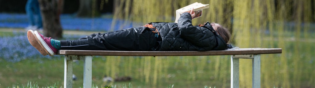 Eine Frau liegt im Stadtpark in Regensburg (Bayern) auf einer Bank und liest ein Buch.