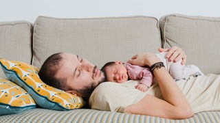 Junger Vater schläft auf dem Sofa und hält ebenfalls schlafenden Säugling auf dem Bauch