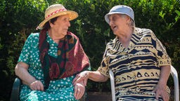 Zwei ältere Frauen mit Sonnehüten sitzen auf Stühlen und unterhalten sich