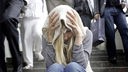 Junge Frau sitzt verloren mit einem Schal über dem Kopf alleine auf einer Treppe, während Menschen an ihr vorbeigehen
