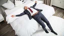 Ein Mann im Anzug liegt die Arme austreckend auf einem Bett.