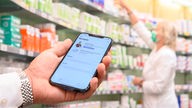 Auf einem Smartphone ist die geöffnete App "Das E-Rezept" zu sehen, während im Hintergrund eine Apothekerin in einer Apotheke an einem Regal steht