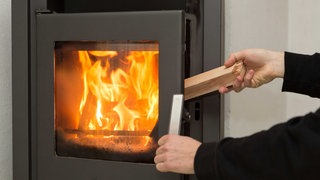 Ein Mann legt in seiner Wohnung ein Stück Holz in einen Kaminofen, in dem ein Feuer brennt.