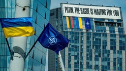 NATO-Gipfel in Vilnius 2023: Eine ukrainische Flagge hängt in Vlinius neben einer NATO-Flagge, an einem Bürogebäude hängt eine Banner: "Putin, the Hague is waiting for you."