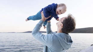 Vater hält am Meer sein Kind lachend mit den Armen in Höhe. 