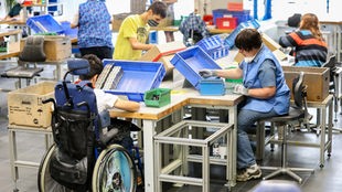 Ein Mann im Rollstuhl montiert mit anderen Mitarbeiterinnen und Mitarbeitern Halterungen für Jalousien in einer Werkstatt. 