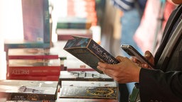 Frau steht vor einem Bücherstapel und hält ein Buch in der Hand. 