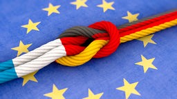 Zwei Seile mit Knoten in den Farben der Flaggen von Frankreich und Deutschland liegen auf der EU-Flagge. 