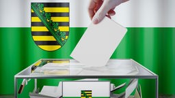 Hand wirft Stimmzettel in eine Wahlurne, im Hinergrund ist die Flagge Sachsens zu sehen. 