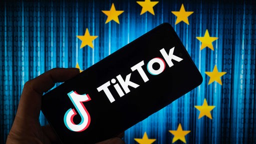 Das TikTok-Symbol wird auf einem Telefonbildschirm angezeigt, im Hintergrund sind in dieser Fotoillustration die Sterne der Europäischen Union zu sehen, 07.08.2023.