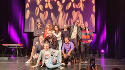 Alle Künstler:innen der Show Köln lacht und Moderator Tobias Mann posieren auf der Bühne im Gloria Theater