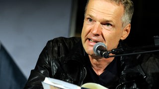 Der Schriftsteller Wolf Haas hält bei einer Lesung ein Buch in d er Hand und spricht in ein Mikrofon