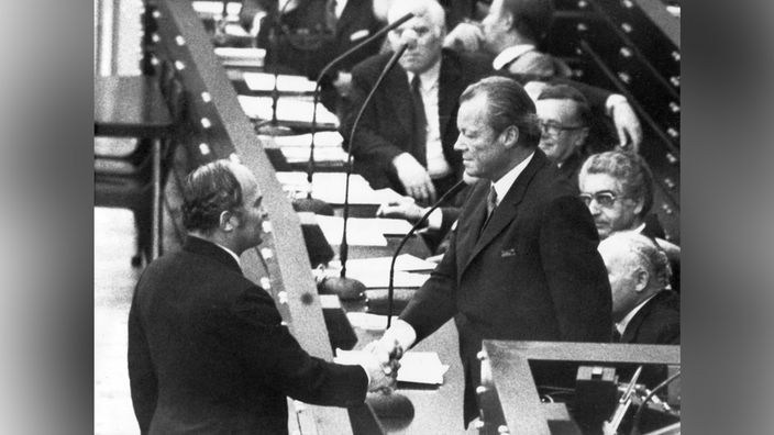 Im Bonner Bundestag reicht Rainer Barzel Bundeskanzler Willy Brandt die Hand, im Hintergrund ist die Regierungsbank zu sehen mit diversen Personen