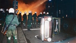 Polizeibeamte bekaempfen am Abend des 24. August 1992 in Rostock-Lichtenhagen eine brennende Strassenblockade mit einem Wasserwerfer. Im Vordergrund liegt ein Auto auf der Seite