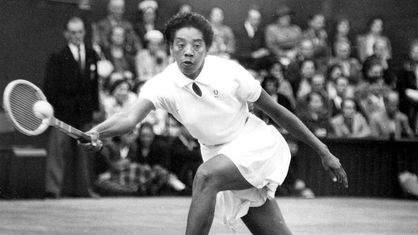 Die Tennisspielerin Althea Gibson in weißem Sportdress streckt sich zur Seite um eine dynamische Vorhand zu spielen, im Hintergrund Publikum (schwarz-weiß-Fotografie)