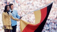 Heidi Schüller spricht als erste Frau überhaupt den olympischen Eid bei der Eröffnungsfeier der Spiele in München, neben ihr steht ein Mann, beide halten eine große Deutschlandfahne