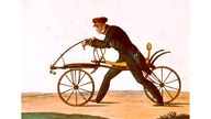 Eine kolorierte Radierung zeigt einen Mann auf dem Laufrad, das Karl Freiherr von Drais zum Patent anmeldete, das Gefährt ist aus Holz zwei Räder werden durch einen geschwungenen Rahmen verbunden