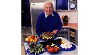 Robert Atkins, ein schlanker Mann mit weißen Haaren steht in einer KÜche und hält freundlich lächelnd einen Teller mit viel Fleisch in die Kamera, Aufnahme aus dem Jahr 1999