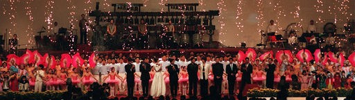 Festlich gekleidete Menschen stehen auf einer Bühne, im Hintergrund bildet ein Feuerwerk einen Vorhang aus Funken