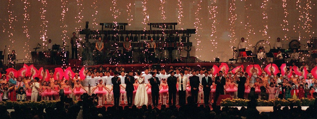 Festlich gekleidete Menschen stehen auf einer Bühne, im Hintergrund bildet ein Feuerwerk einen Vorhang aus Funken