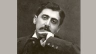Schwarz-Weiß-Fotografie mit Sepiatönen zeigt Marcel Proust. Er sitzt auf einem Stuhl, neigt den in eine Hand gestützen Kopf. Er trägt einen Schnurrbart, hat volles, dunkles Haar.