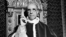Papst Pius XII. hebt die rechte Hand mit dem Ring des heiligen Petrus, um einen Segen zu sprechen.