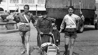 Zwei Schülerlotsen haben eine Frau mit Kinderwagen und einem kleinen Jungen in die Mitte genommen und führen sie mit erhobenen Kellen über eine Straße (Aufahme von 1953)