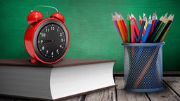 Sybmbolbild für eine Schulstunde: Ein Wecker steht auf einem Buch, daneben eine Dose mit Buntstiften.