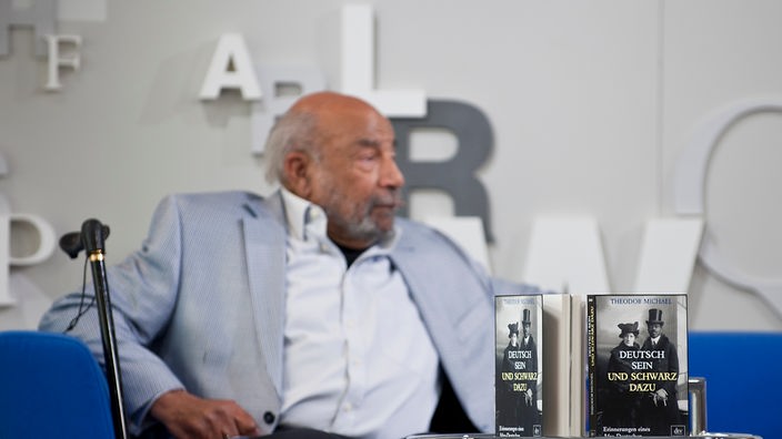 Theodor Michael sitz auf einer blauen Couch links sein Gehstock, vor ihm auf dem Tisch sein Buch "deutsch sein und schwarz sein "