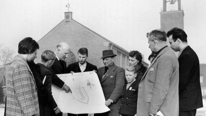 Lagerpfarrer Wilhelm Scheperjans (3.v.l.) zeigt Flüchtlingen aus Böseckendorf am 23.02.1963 im Lager Friedland den Bauplan für die geplante Neubausiedlung Neu-Böseckendorf