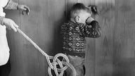 Frau schlägt Kind mit Teppichklopfer (gestelltes Foto aus den 1950er Jahren)