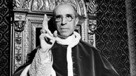 Papst Pius XII. hebt die rechte Hand mit dem Ring des heiligen Petrus, um einen Segen zu sprechen.