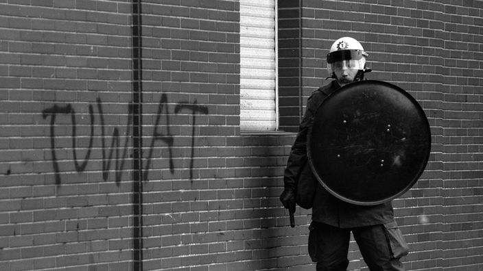 Polizist mit Schutzschild, Helm und Schlagstock an einer Wand mit dem Spruch "Tuwat" (Tue was - Anspielung auf den "Tunix"-Kongress von 1978 in der TU-Berlin) bei einer Demonstration 1981 beim Besuch des US-Verteidigungsministers Alexander Haig in Berlin