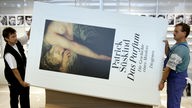 Zwei Monteure tragen das überdimensionale Buch "Das Parfum" von Patrick Süskind durch eine Halle. 