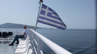 Christian Dassel sitzt auf einem Boot in Griechenland, neben ihm weht die griechische Flagge im Wind